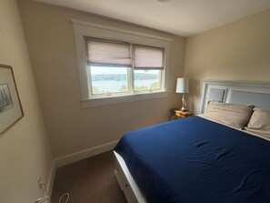 A bedroom with a bed, a nightstand, a lamp, a window with blinds, and a framed picture on the wall.
