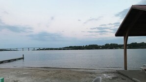 A sandy beach with a view of a calm body of water, a bridge in the distance, and a covered seating area with chairs.