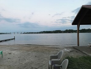 A sandy beach with a view of a calm body of water, a bridge in the distance, and a covered seating area with chairs.