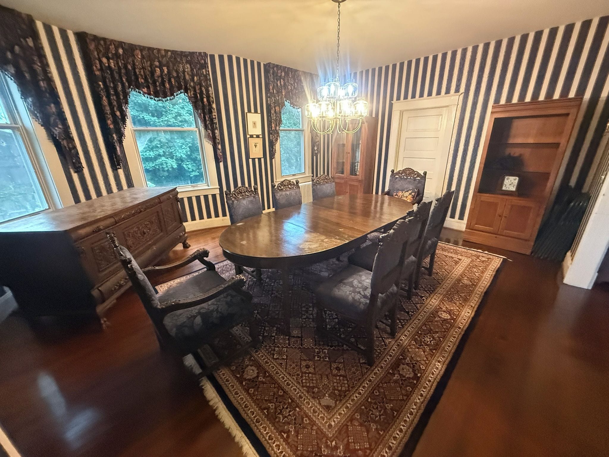 Ein Esszimmer mit einem runden Tisch, Stühlen, einer Chandelieber und gestreifter Tapete.