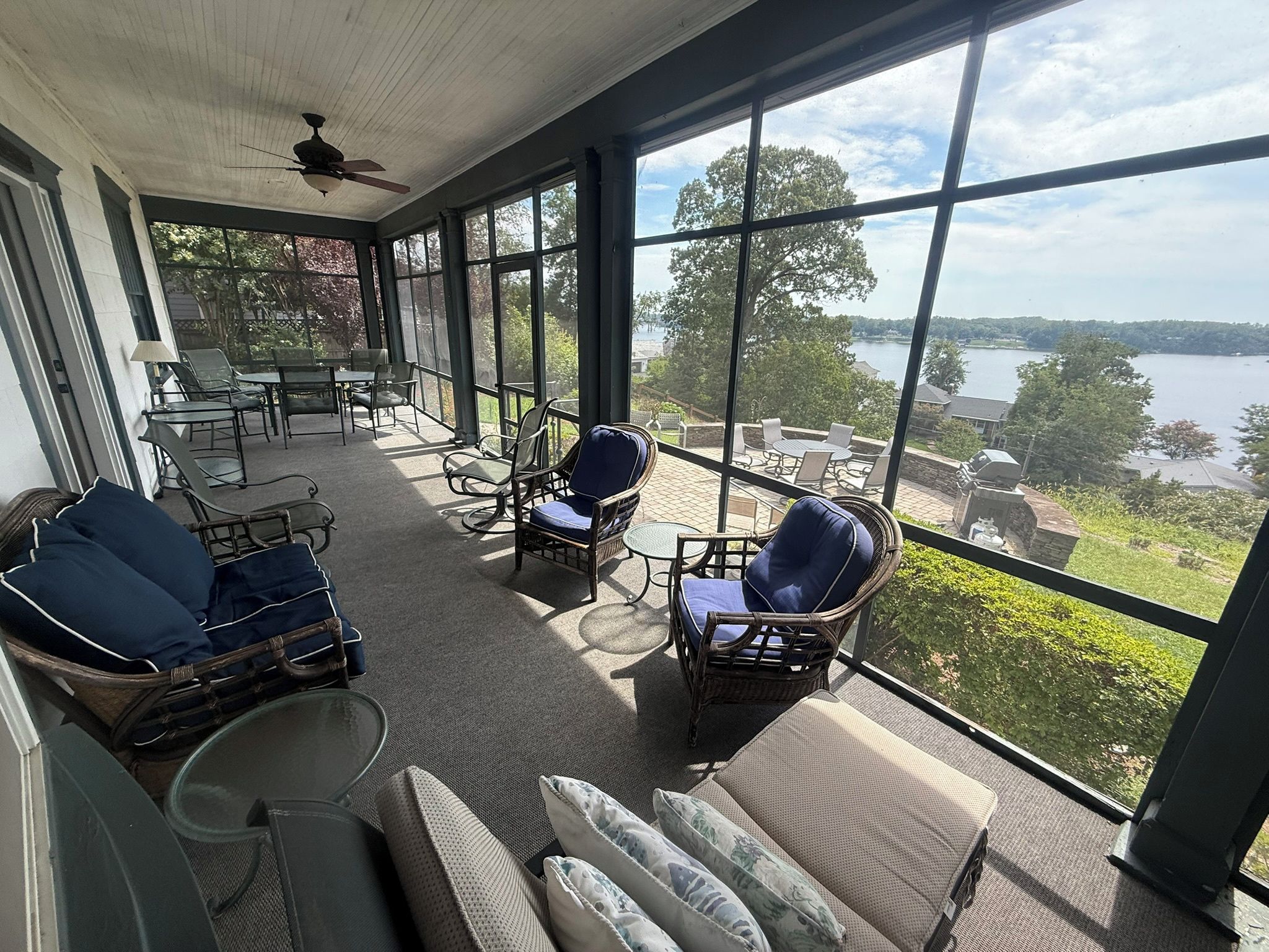 Une terrasse couverte, meublée de meubles en osier, dotée d’un ventilateur de plafond et offrant une vue sur un lac.