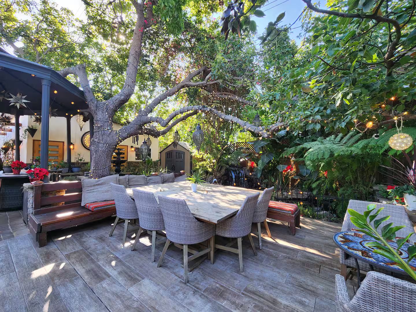 An outdoor patio with a wooden table and wicker chairs, surrounded by lush greenery and a gazebo.