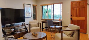 A living room with a television, a desk, two chairs, and a window with blinds. - Cozy 2BR on the Jackson Market Courtyard (Culver City)