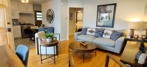 Ein Wohnzimmer mit einer Couch, einem Couchtisch und Sesseln, ein Essbereich mit Tisch und Stühlen sowie eine Küche im Hintergrund.