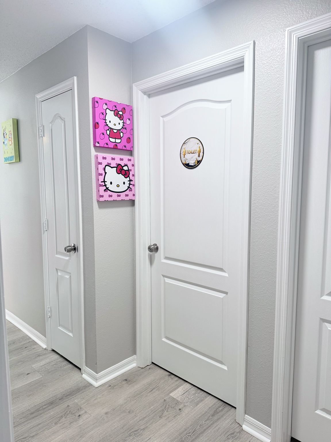Un couloir avec des portes blanches, dont une ornée d’un autocollant Hello Kitty et une autre portant un panneau indiquant les toilettes.