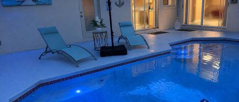 An indoor swimming pool with two lounge chairs and a table, a ceiling fan, and a large umbrella.