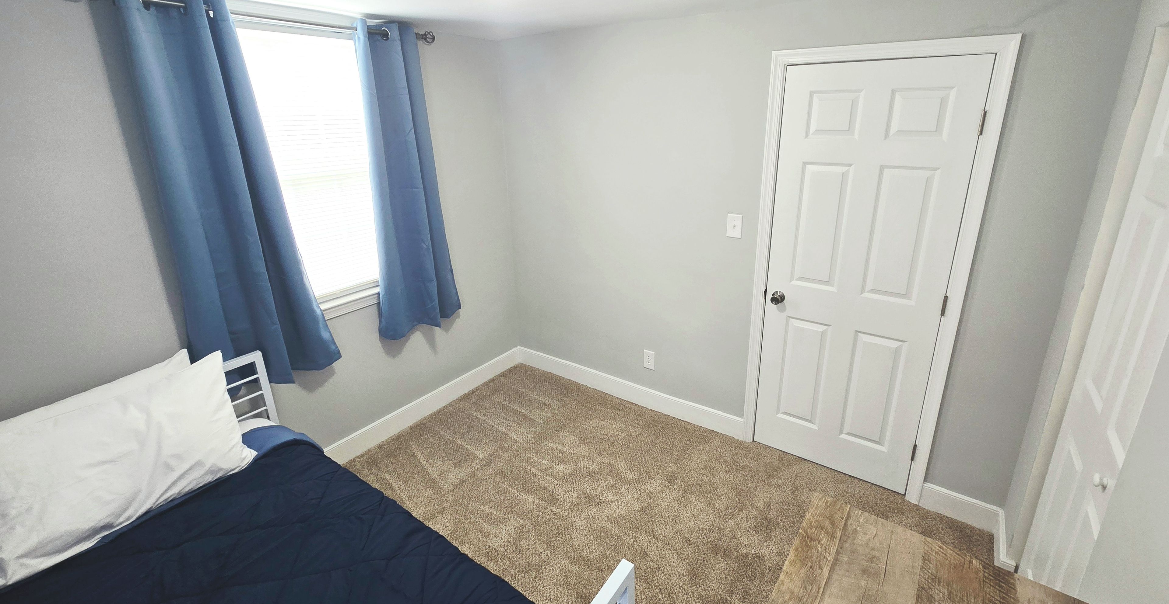 A bedroom with a bed, a window with curtains, a door, and a carpeted floor.
