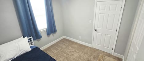 A bedroom with a bed, a window with curtains, a door, and a carpeted floor.