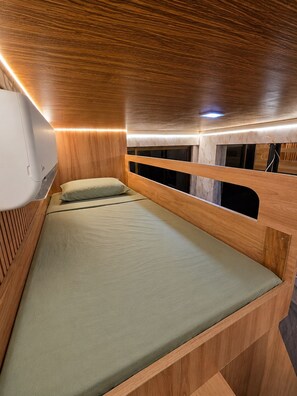 A compact, wooden bunk bed with a single mattress and pillows, a small window, and built-in lighting.