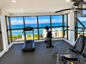 A gym with exercise equipment and large windows overlooking a beach.