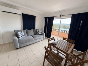 Ein Wohnzimmer mit grauem Sofa, gemusterten Kissen, einem Holztisch und Stühlen. Ein großes Fenster mit Vorhängen, und eine Klimaanlage an der Wand.