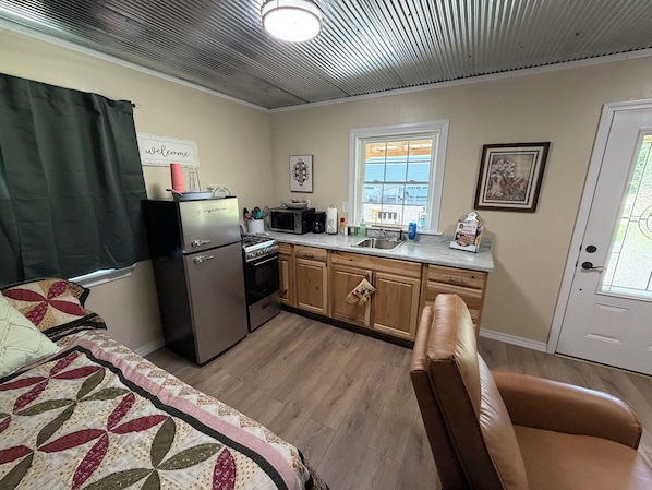 A compact kitchen with a refrigerator, stove, microwave, and sink. There is a window with a view of the outside, a door, and a bed with a quilt.