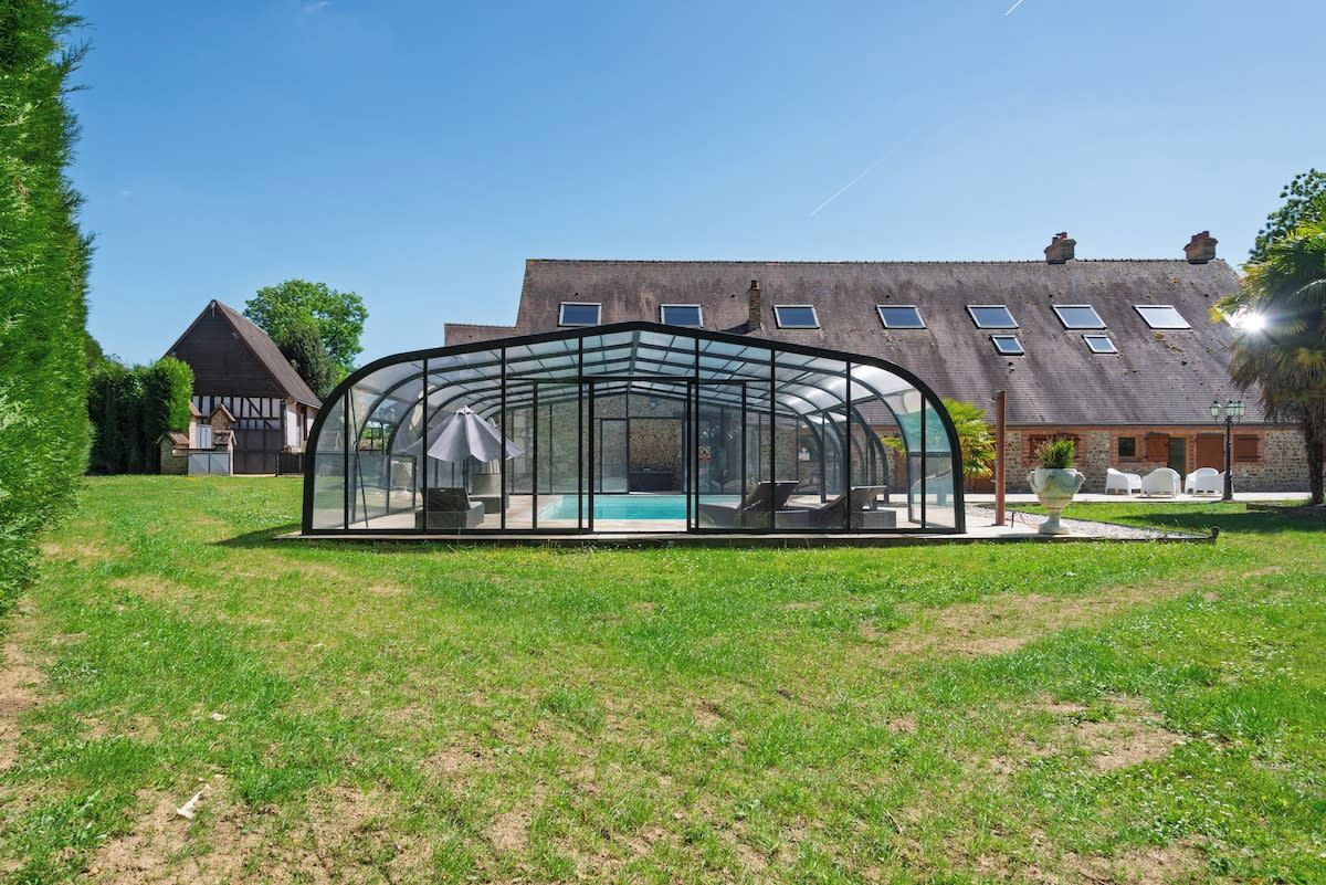 A glass-enclosed outdoor area with a pool, surrounded by grass and a building with a sloped roof.