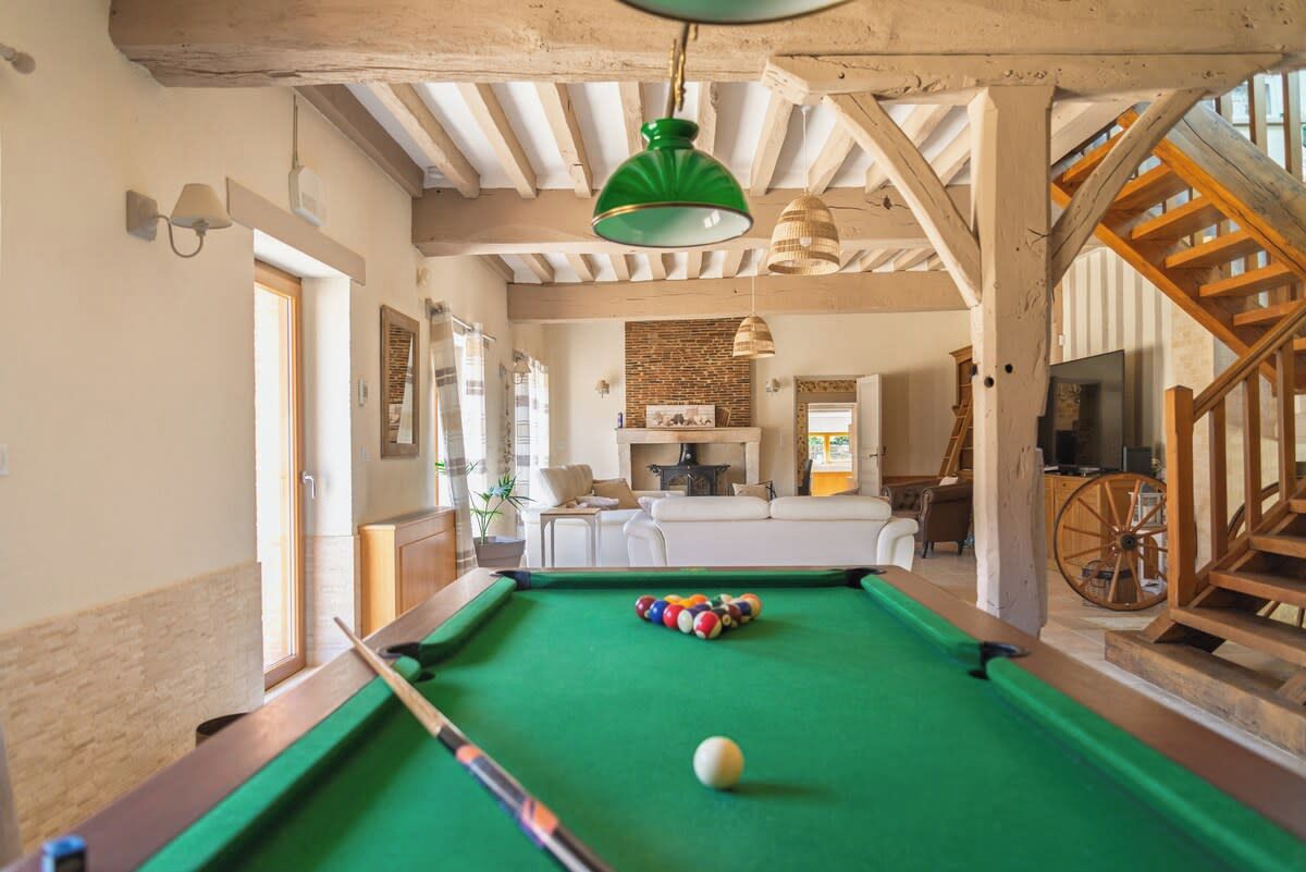 Une table de billard dans une pièce aux poutres apparentes, avec un luminaire vert suspendu et une esthétique rustique.