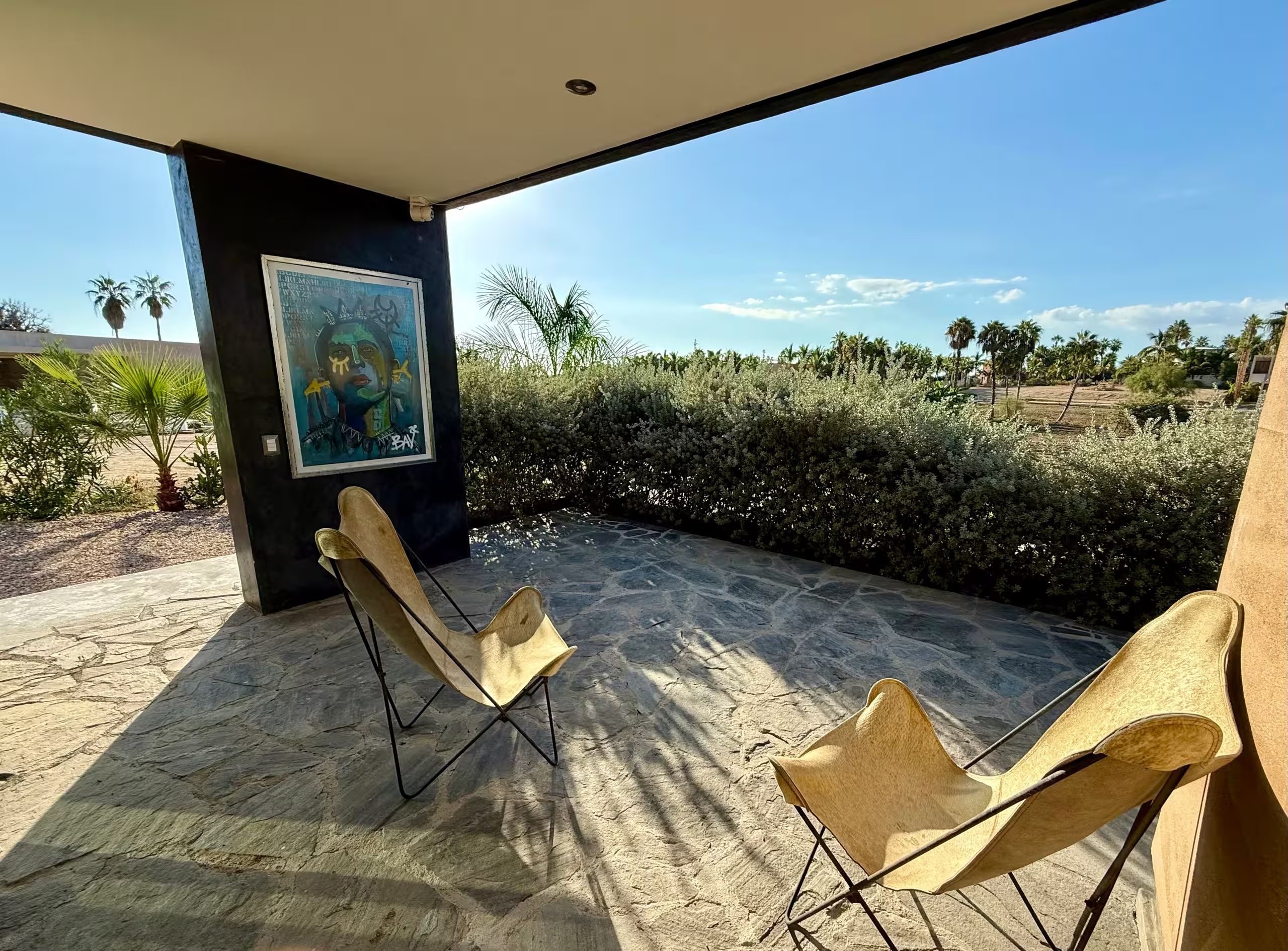 Eine Außenpatio mit Steinboden, zwei Klappstühlen, einem großen abstrakten Gemälde an einer Wand und Blick auf eine angelegte Grünfläche mit Bäumen und Sträuchern.