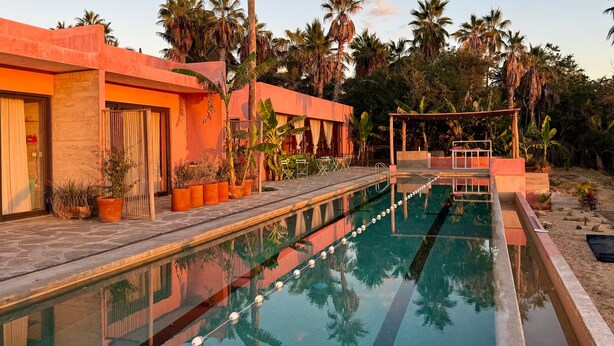 Piscina com deck de madeira, rodeada por palmeiras e um edifício com fachada laranja.