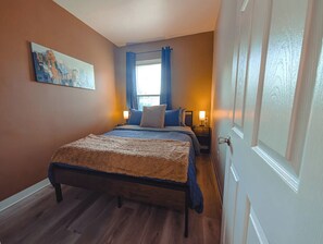 A bedroom with a bed, a nightstand, a lamp, a window with curtains, and a painting on the wall. - Historic Detroit Charm • 10 min Downtown (Detroit)