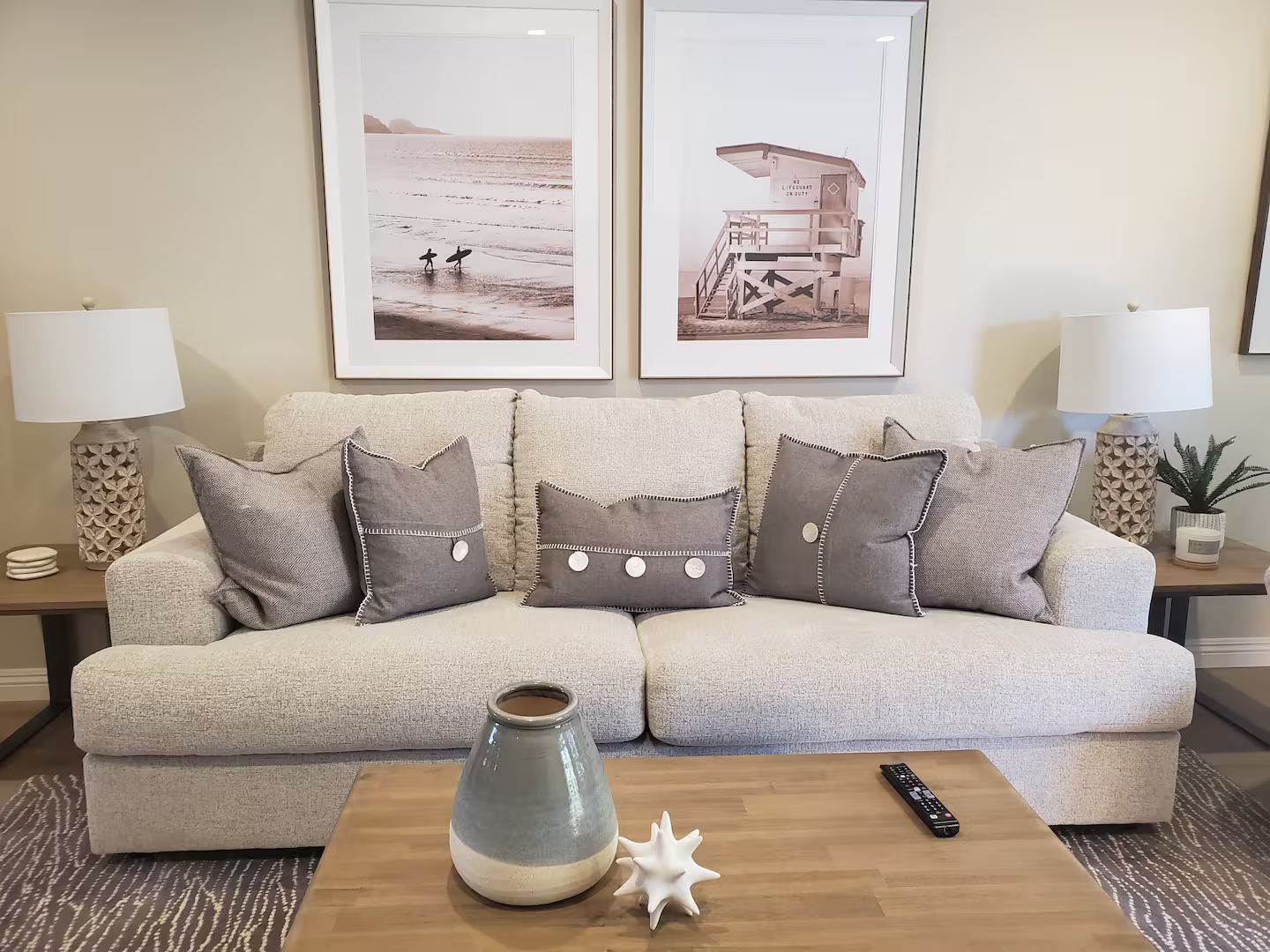 Un salon avec un canapé beige, des coussins décoratifs, un vase et deux photographies de plage encadrées accrochées au mur.