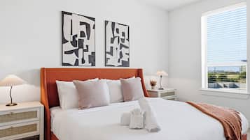 Une chambre moderne avec un grand lit, deux tables de chevet, une fenêtre avec des stores et des œuvres d’art abstraites accrochées au mur.