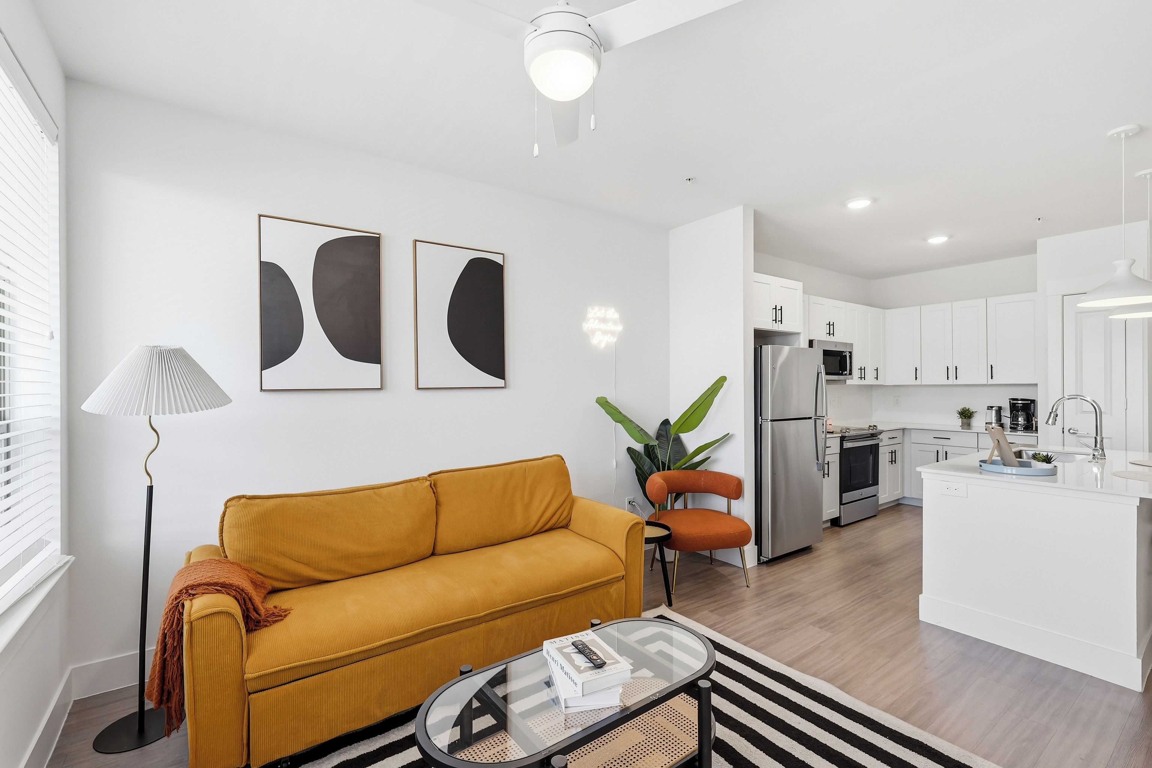 Una sala moderna con un sofá, una mesita de centro y una lámpara. Hay una zona de cocina con armarios y electrodomésticos.