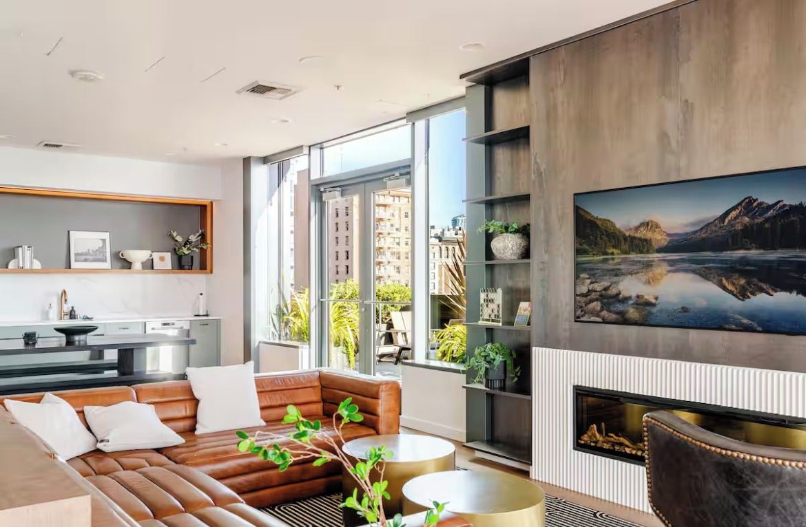 Uma sala de estar moderna com sofá, mesa de centro e lareira. Há uma grande janela que oferece vista da cidade.