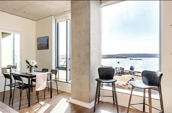 A dining area with a table and chairs, a large window with a view of a waterfront, and a concrete pillar.