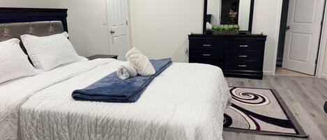 A neatly made bed with white bedding and a blue towel, a dark wooden dresser with a mirror, and a patterned rug on a wooden floor.