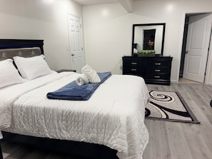 Cama bem arrumada com roupa de cama branca e toalha azul, cômoda de madeira escura com espelho e tapete estampado sobre piso de madeira.