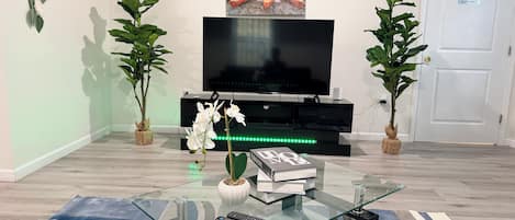 Una sala de estar con una mesa de centro de vidrio, un televisor de pantalla plana, dos plantas en macetas y un cuadro en la pared.