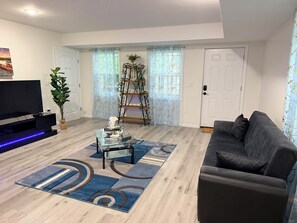 Ein Wohnzimmer mit einem Flachbildfernseher, einem Glastisch, einem grauen Sofa und einem gemusterten Teppich.