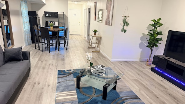 A modern living room with a glass coffee table, a sofa, a dining area with chairs, a TV, and a plant.