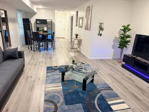 A modern living room with a glass coffee table, a sofa, a dining area with chairs, a TV, and a plant.