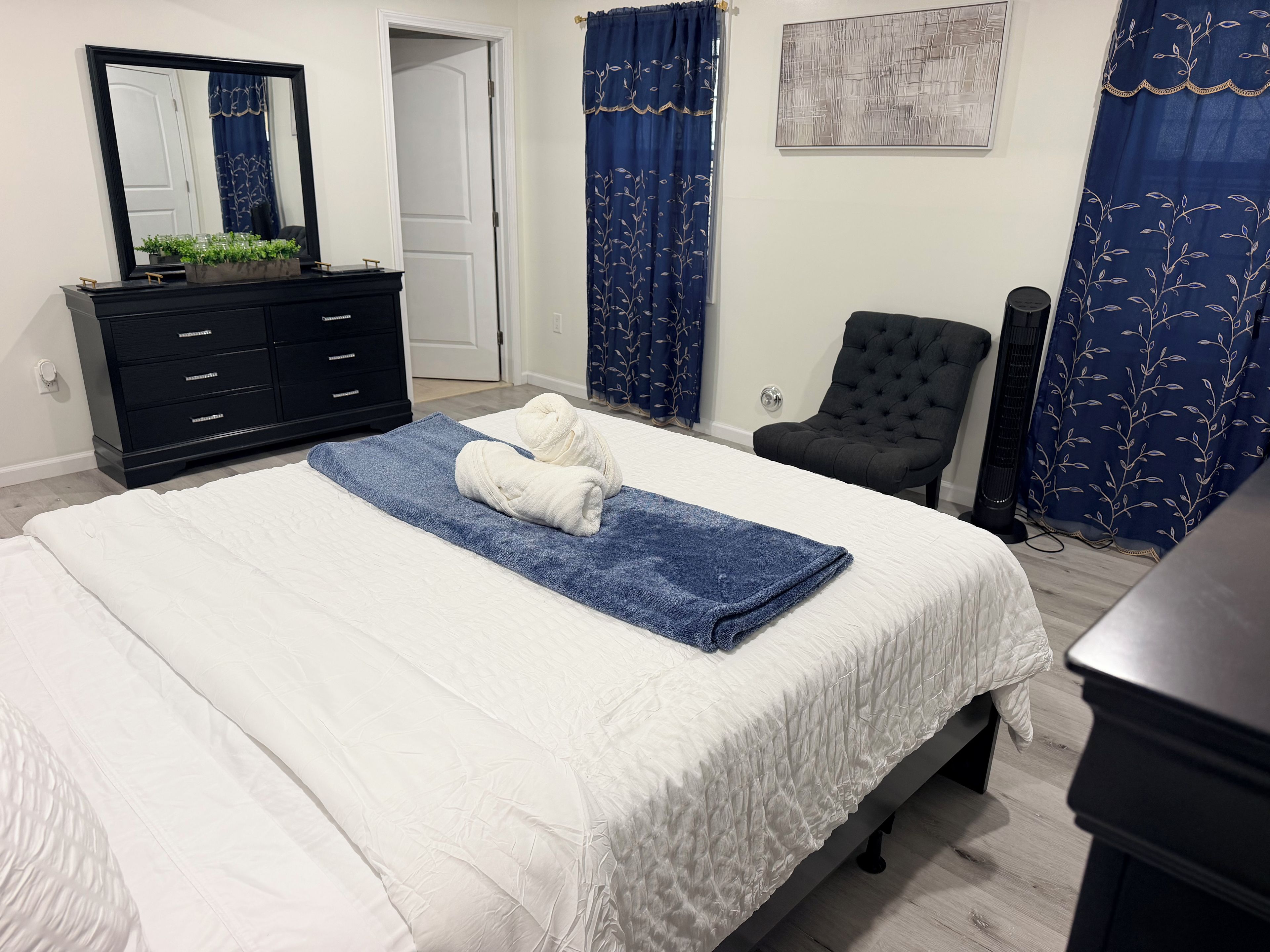 Ein ordentlich bezogenes Bett mit einem blauen Handtuch und zwei weißen Handtüchern, eine schwarze Kommode mit Spiegel, ein schwarzer Stuhl und ein Standventilator.