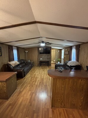 A living room with a wooden floor, a black leather sofa, a wooden coffee table, and a television mounted on the wall. - Welcoming 2-bedroom mobile home with peaceful setting (Eucha)