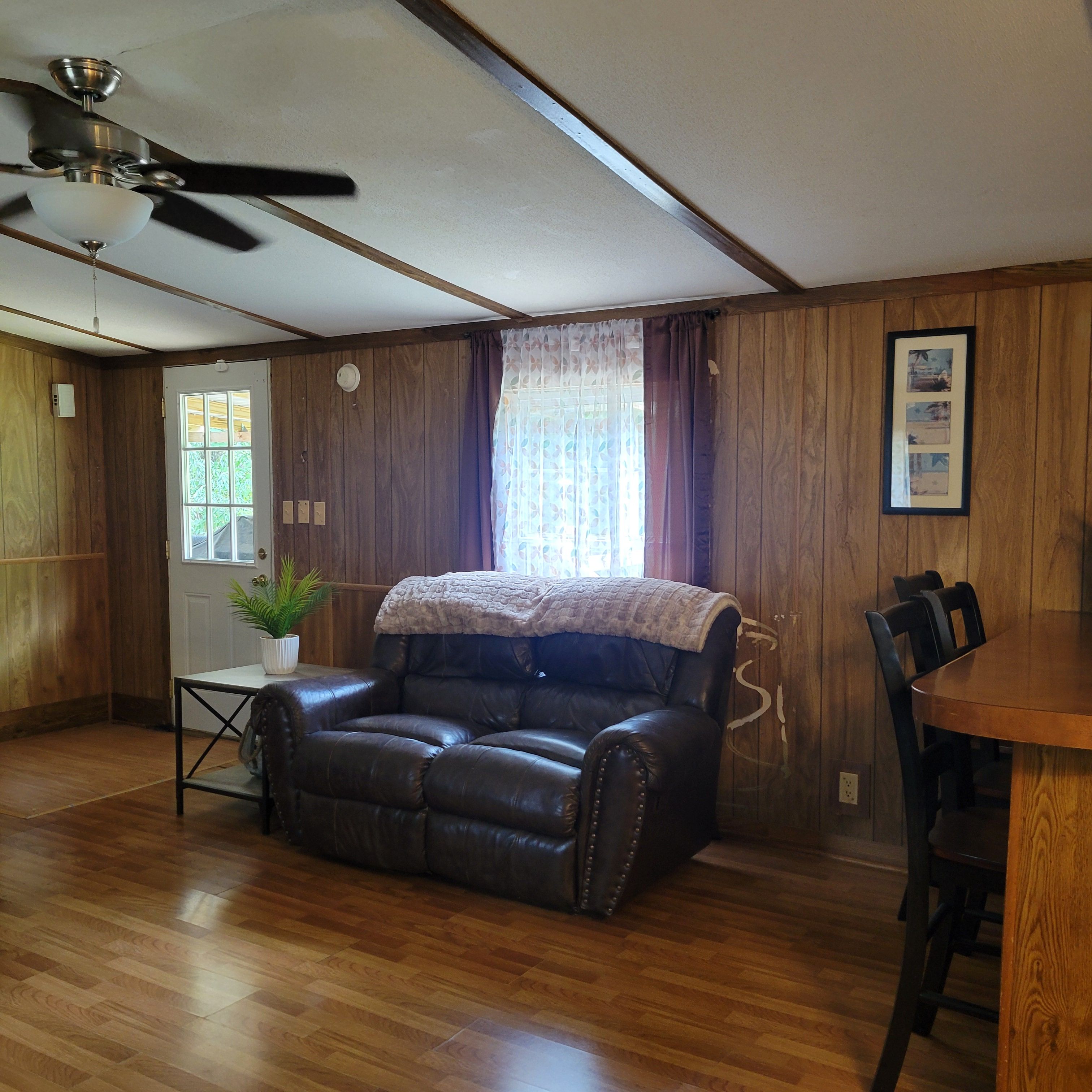 Ein Zimmer mit Ledersofa, Esstisch, Deckenventilator und Holzwänden.