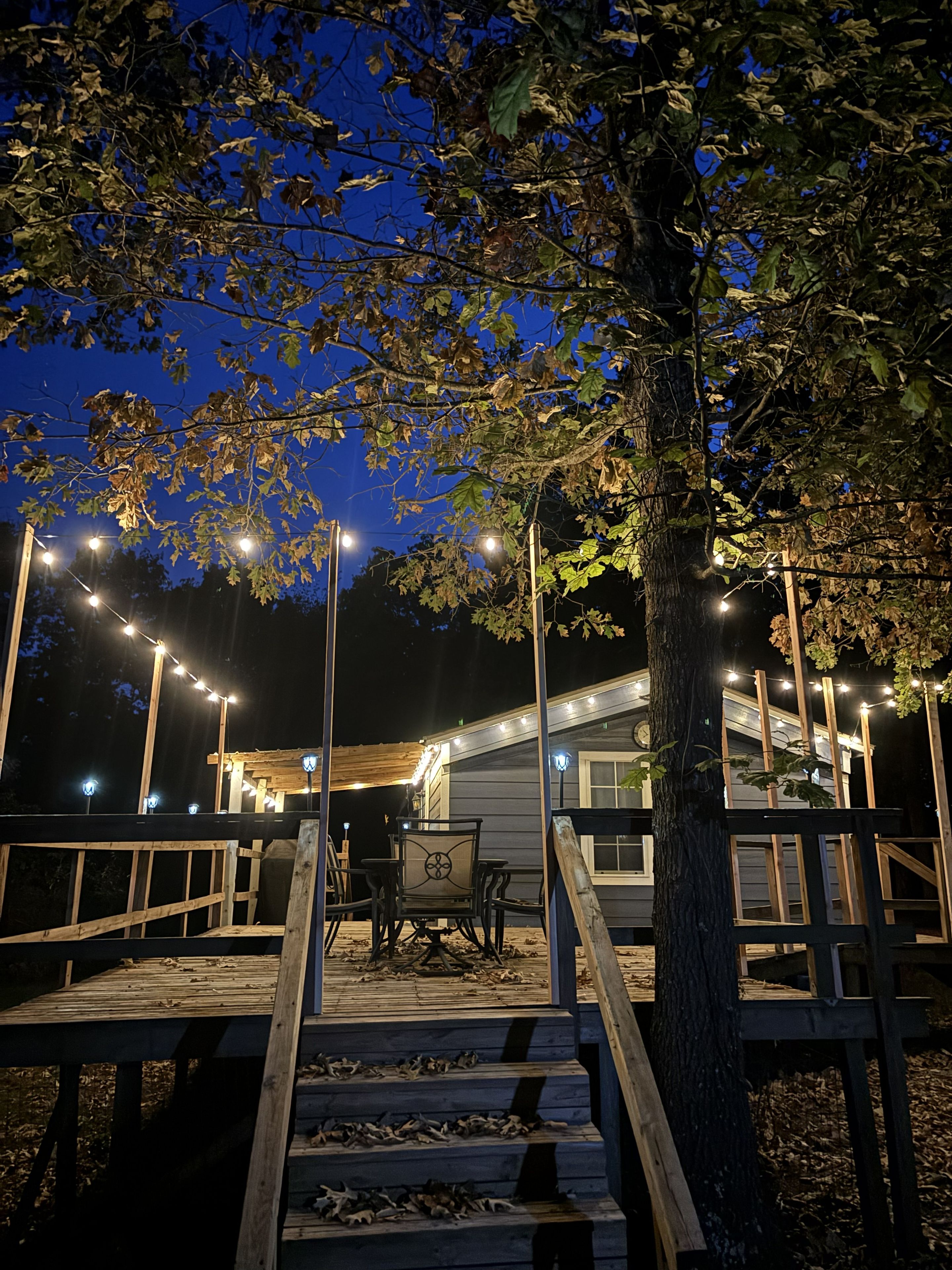 A wooden deck with a staircase leading to a building, surrounded by trees and string lights.