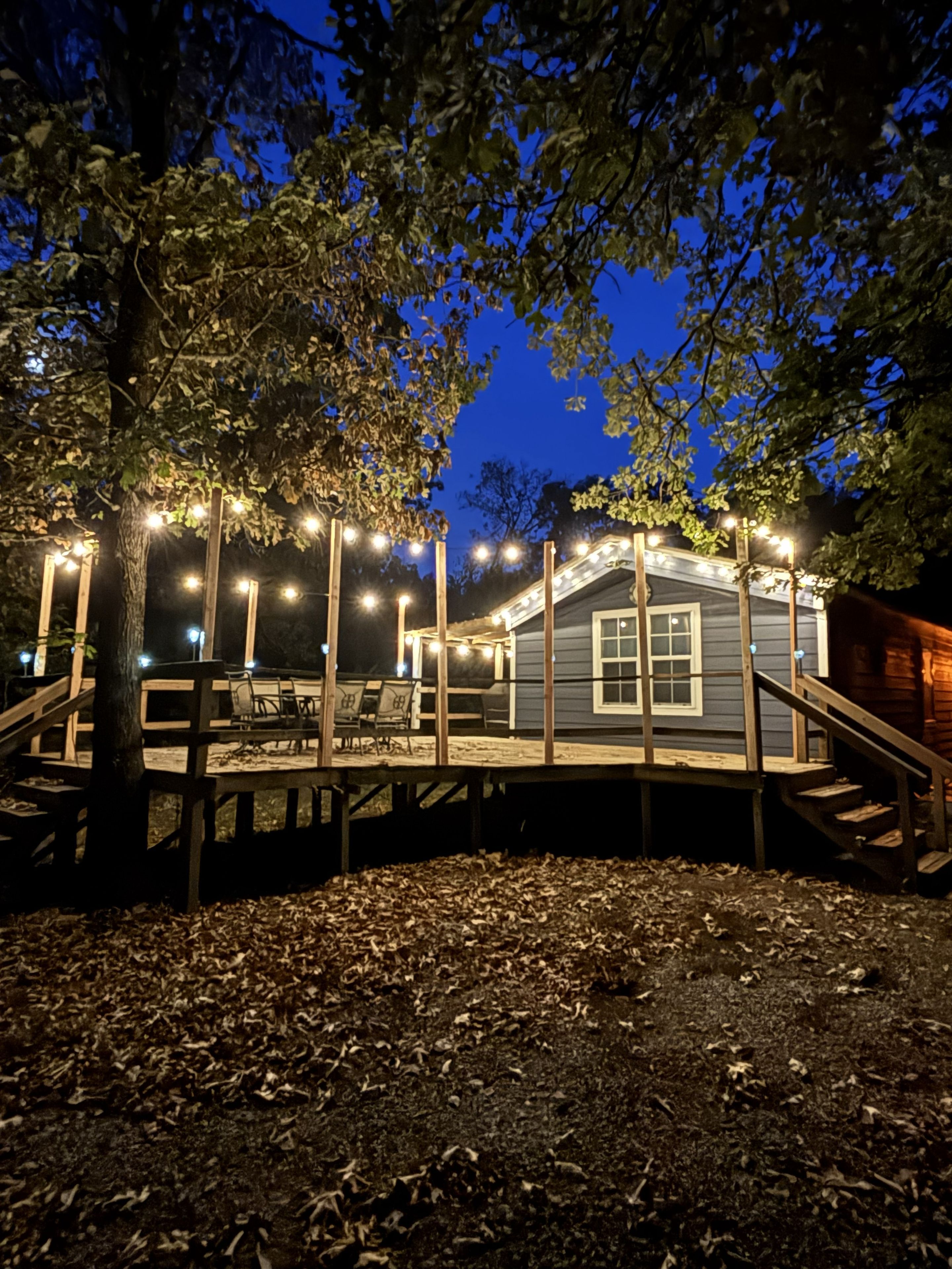 A wooden deck with a staircase leading to a building, surrounded by trees and illuminated by string lights.
