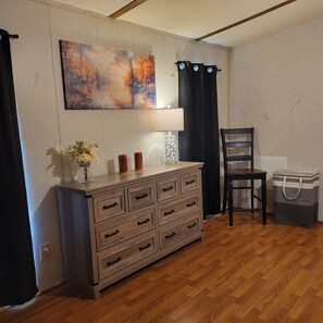 A room with a wooden dresser, a chair, a lamp, a trash bin, and a painting on the wall. - Welcoming 2-bedroom mobile home with peaceful setting (Eucha)