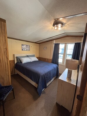 A bedroom with a bed, a bedside table with a lamp, a window with curtains, and a wall clock. - Welcoming 2-bedroom mobile home with peaceful setting (Eucha)