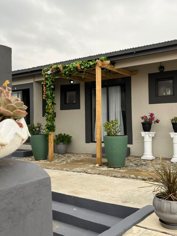 Ein überdachter Außenbereich mit einer Holz-Pergola, Blumentöpfen und einem Betonweg, der zu einem Gebäude mit Fenstern und einer Tür führt.