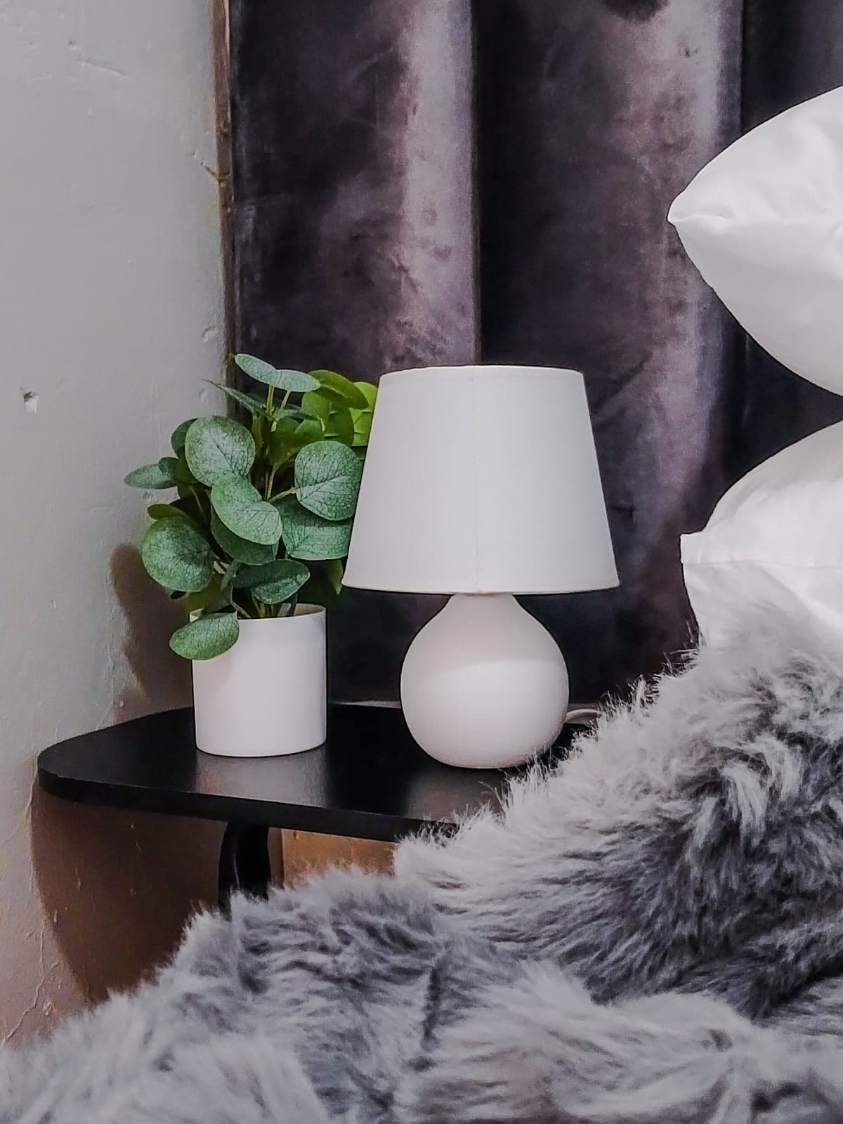 A bed with a white pillow, a white lamp on a black bedside table, and a green plant in a white pot.