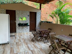 Eine Außenpatio mit Fliesenboden, Holzmöbeln, einem Spülbecken und Blick auf eine rote Wand und Grünpflanzen.