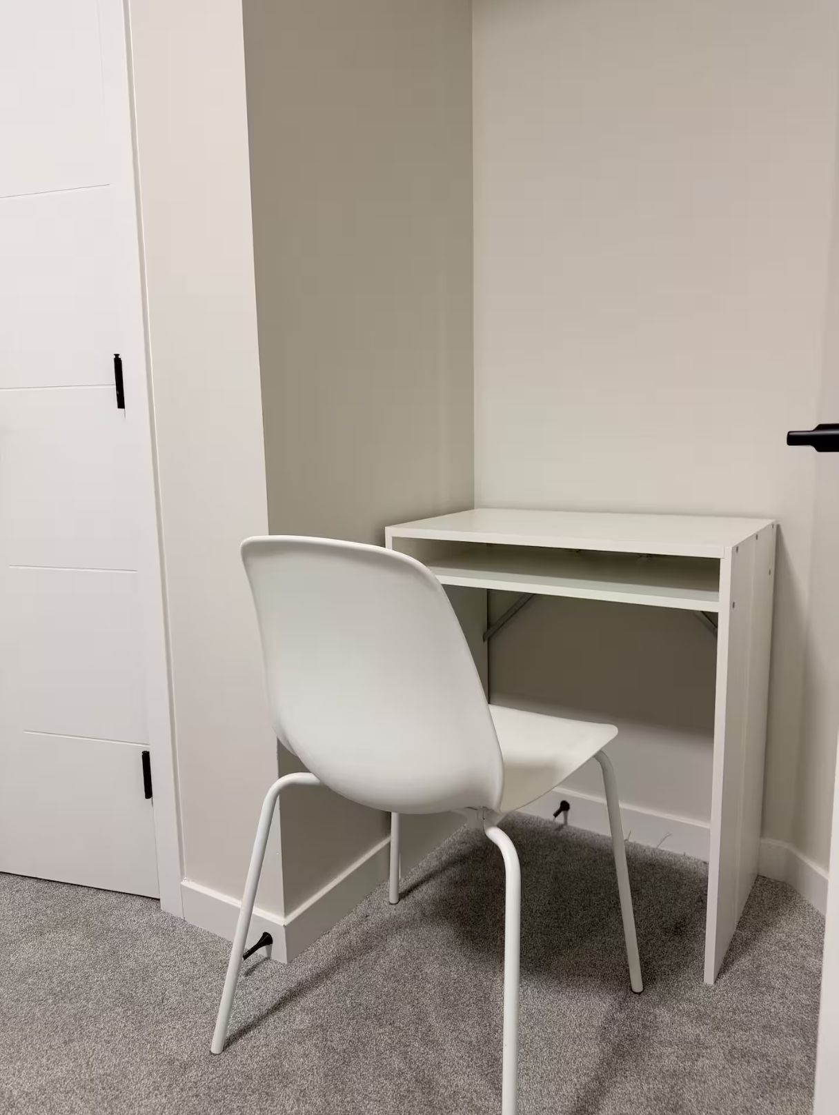 Ein weißer Schreibtisch mit Stuhl steht in einer Ecke des Raumes, der eine Tür und einen Teppichboden hat.