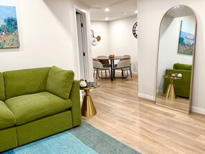 A modern living room with a green sofa, a dining area with a round table and chairs, and a large mirror reflecting the room.