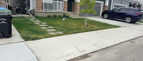 A residential street with houses, a sidewalk, a tree, and a parked car.