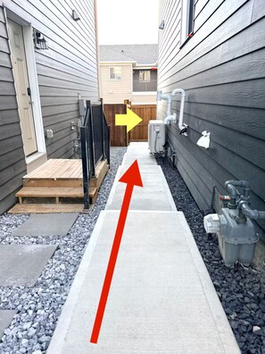 A narrow pathway leading to a wooden deck, flanked by buildings with visible utility pipes and a metal gate. - Beautiful suite close to Spruce Meadows in Calgary, Canada (Calgary)