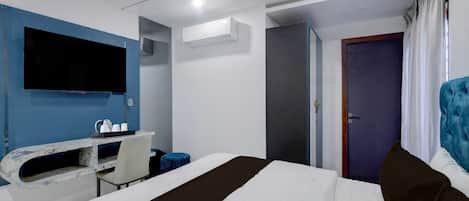 A hotel room with a bed, a desk with a chair, a flat-screen TV mounted on the wall, and a door leading to another room.