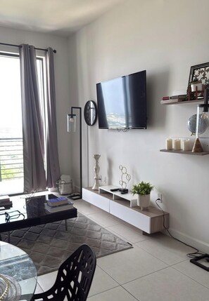 Un saló modern amb televisió de pantalla plana penjada a la paret, una taula de consol blanca amb objectes decoratius, una zona de menjador amb taula de vidre i cadires, i una finestra gran amb cortines.