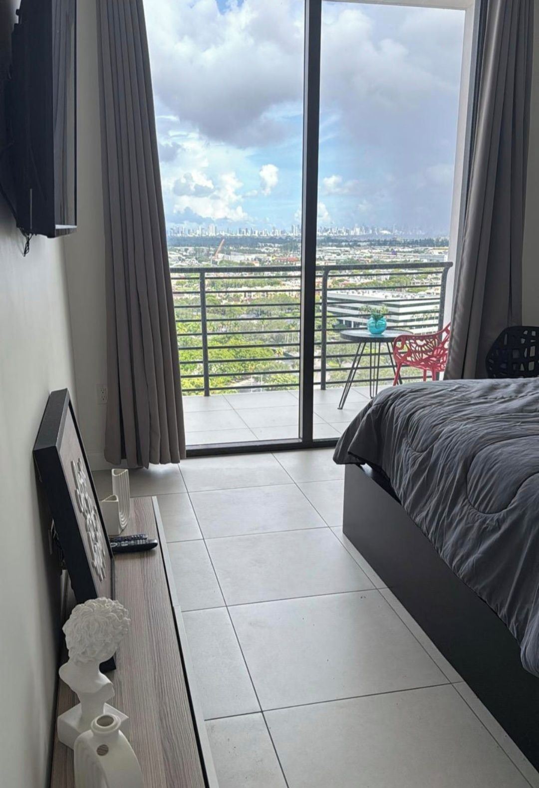 A bedroom with a bed, a TV, a sculpture, and a balcony with a view of a cityscape.