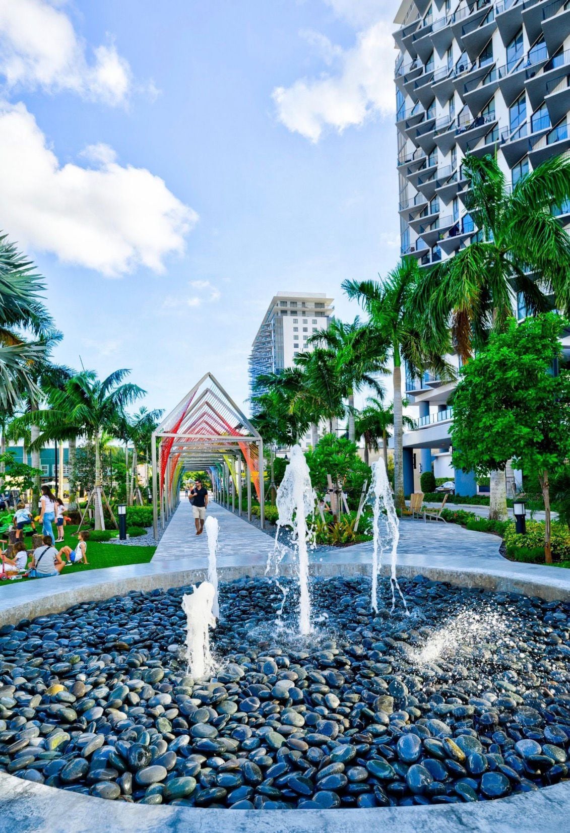 A modern building with a unique geometric facade, a central water feature with multiple fountains, and a pathway lined with palm trees.
